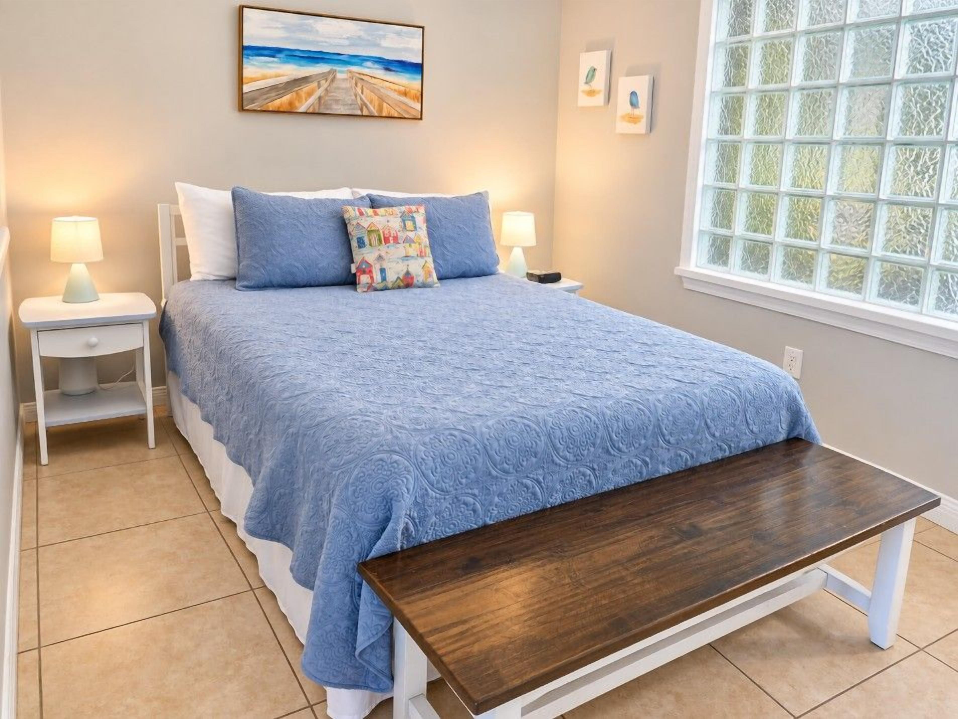 A bedroom with a bed, bedside tables, a bench, a window with frosted glass, and wall art.
