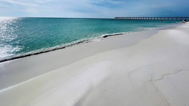 Une plage avec un ciel dégagé, une eau turquoise et une jetée visible au loin.