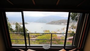 A room with a large window offering a view of a lake and mountains. There are two chairs and a small round table in the room. - Rakuyu (Fujikawaguchiko)