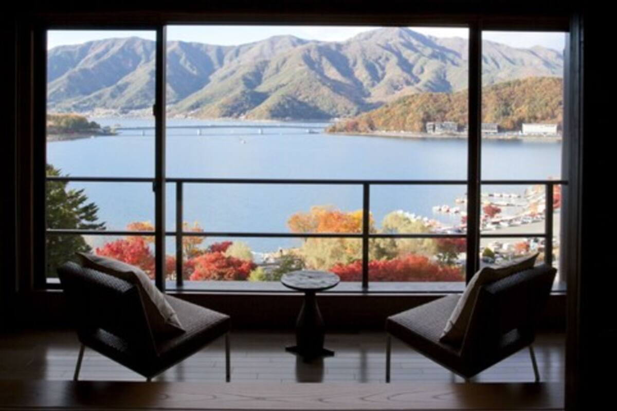 Habitación con un ventanal grande que ofrece vistas a un lago y montañas. Hay dos sillas y una pequeña mesa en la habitación.