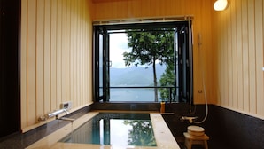 A bathroom with a large bathtub, a toilet, and a window offering a view of a lake and mountains. - Rakuyu (Fujikawaguchiko)