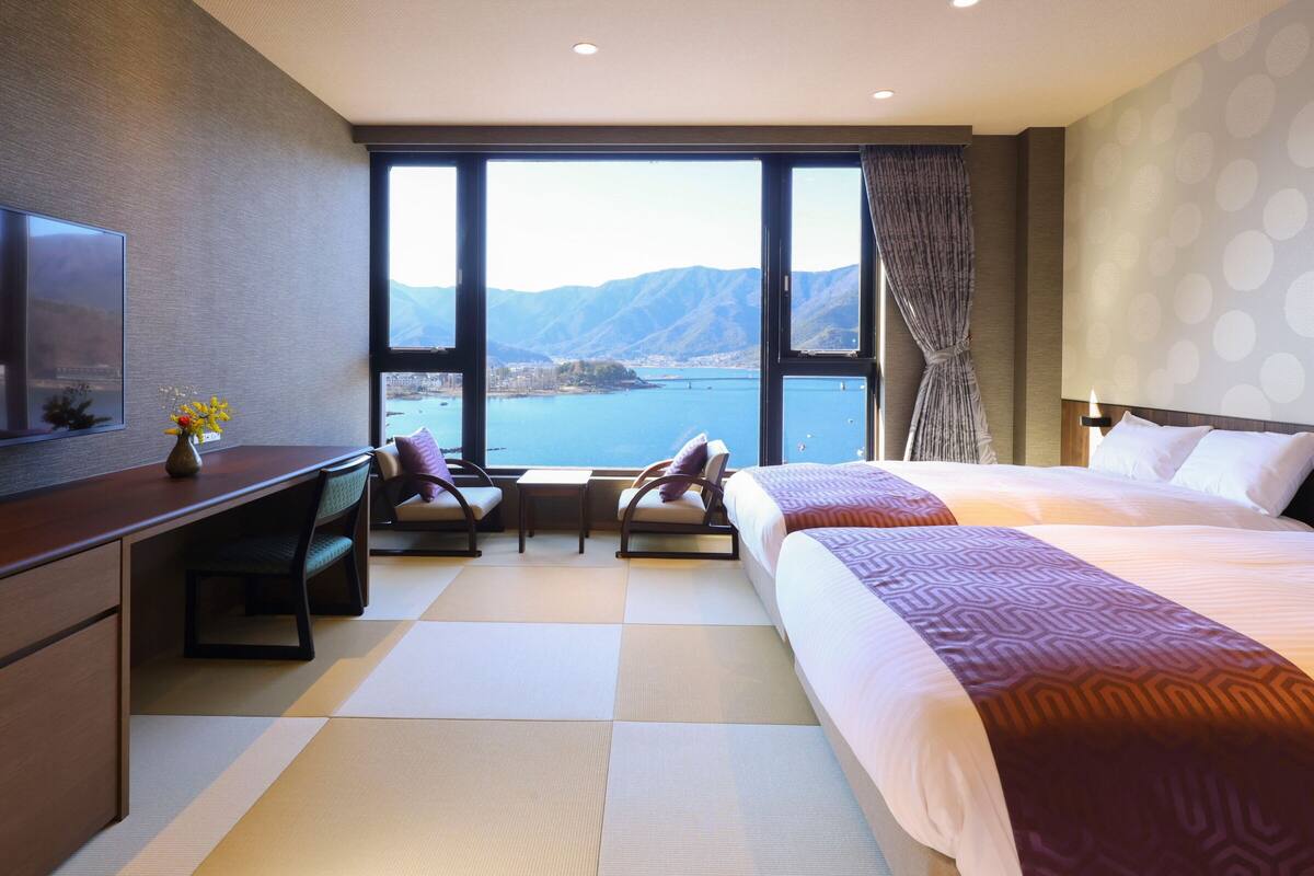 Habitación de hotel con un ventanal amplio que ofrece vistas a un lago y montañas, una cama, un escritorio con silla y un televisor.