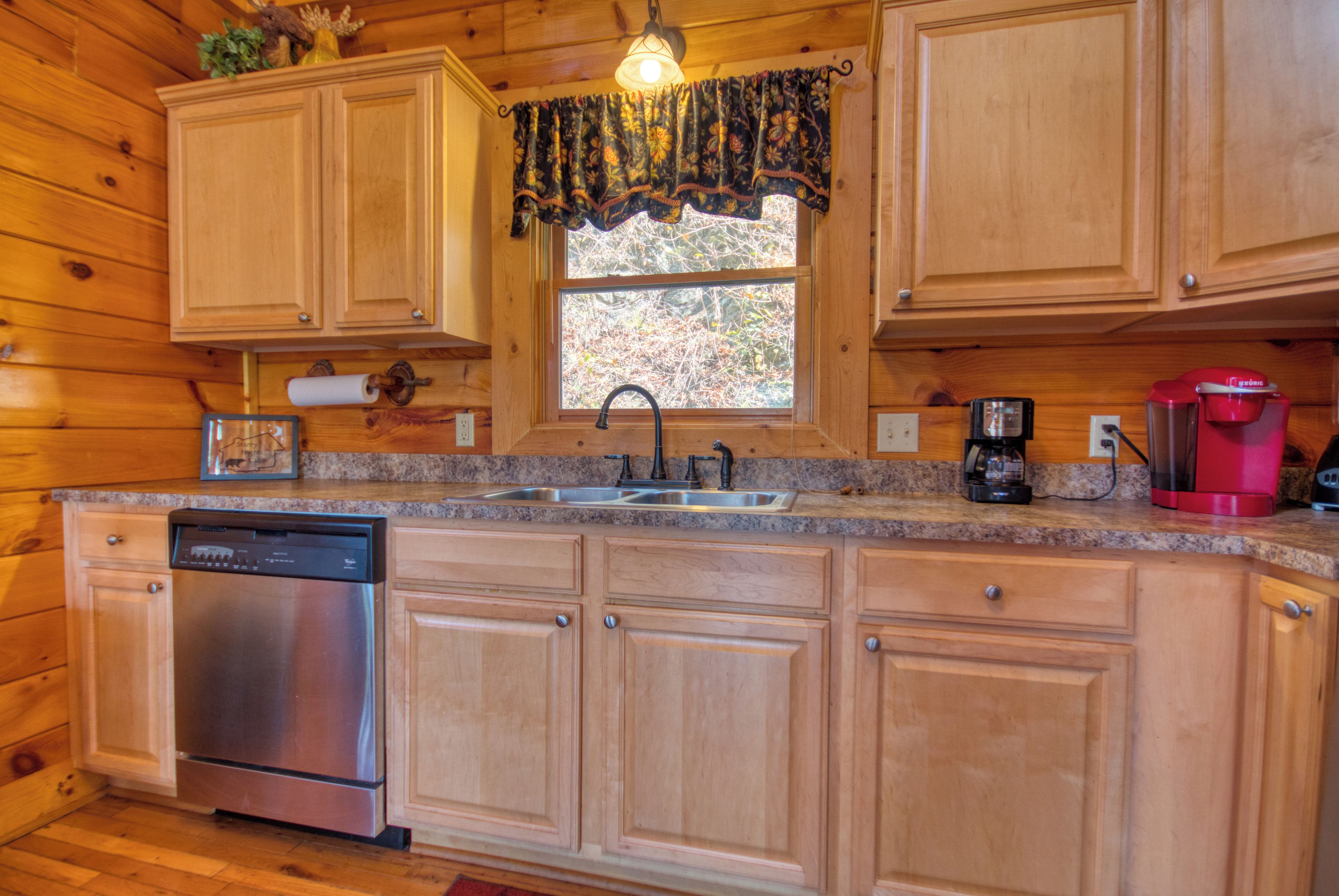 A wooden cabin kitchen with a dishwasher, a sink, a window with a view, and a coffee maker.