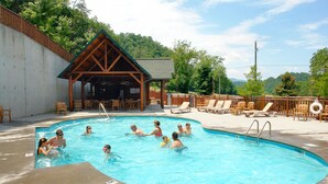 Une piscine où se baignent des gens, une terrasse en bois, des chaises longues et un espace de repos abrité.