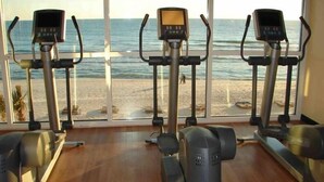 Ein Fitnessraum mit Laufbändern und einem Crosstrainer, mit Blick auf einen Strand.