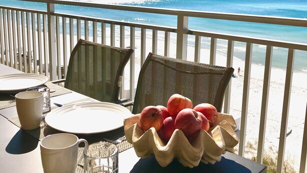 Ein Tisch für zwei Personen mit Obstschale, einem Glas Wasser und Tellern auf einem Balkon mit Blick auf einen Strand.