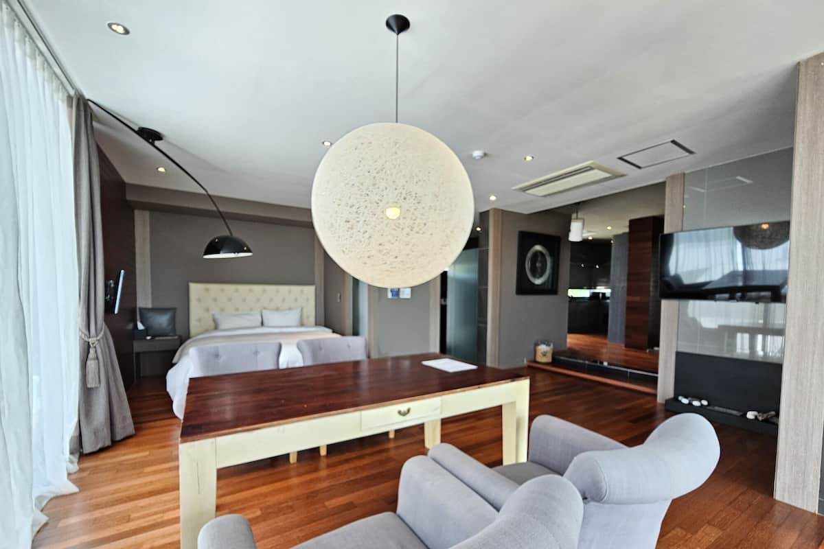 A modern hotel room with a wooden floor, a large bed with a headboard, a dining table with chairs, a large round light fixture, and a TV mounted on the wall.