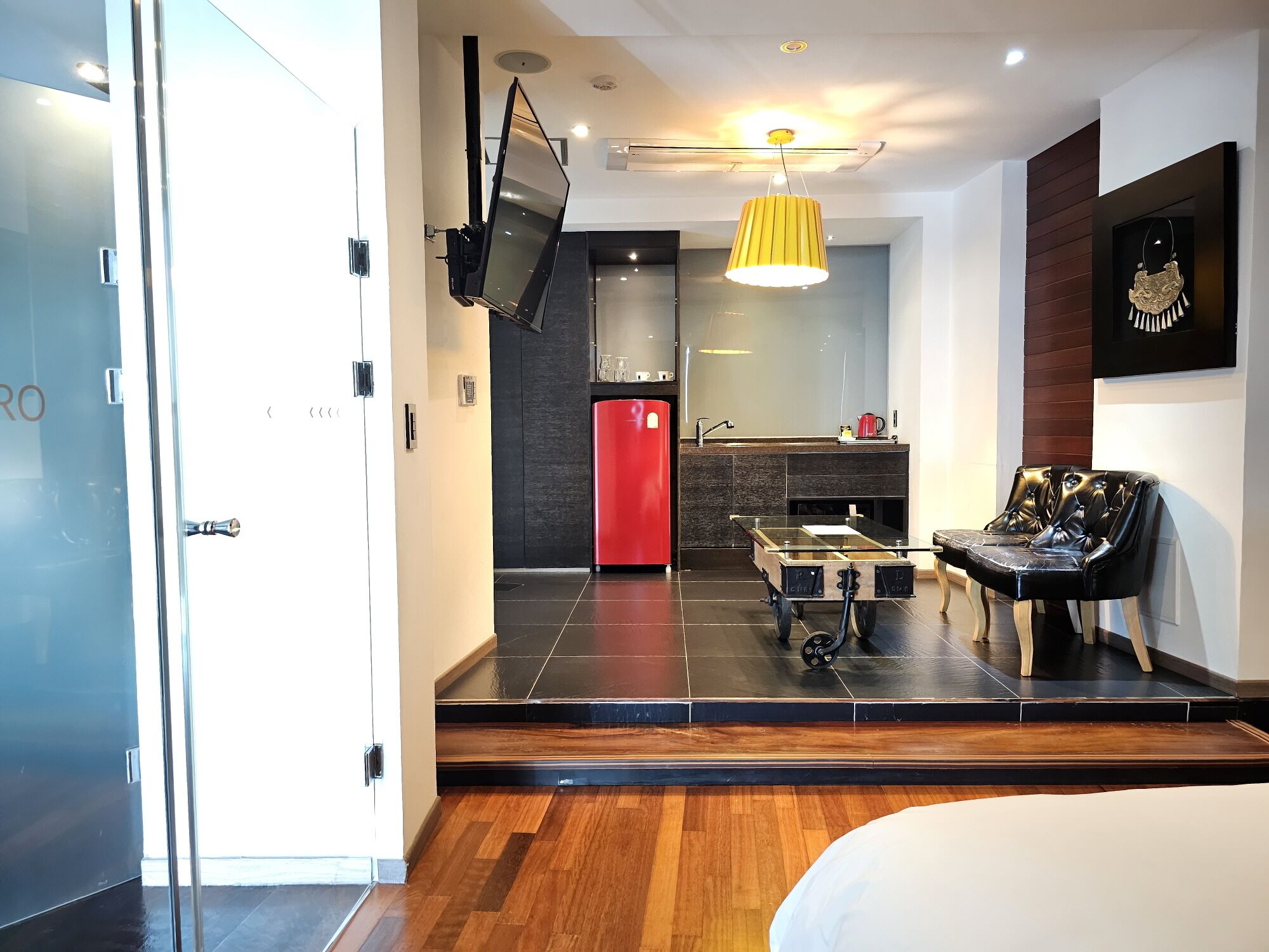 Une chambre d’hôtel moderne dotée d’une porte coulissante, d’un téléviseur à écran plat fixé au mur, d’un réfrigérateur rouge, d’une table à manger en verre avec une chaise noire et d’un plancher en bois.