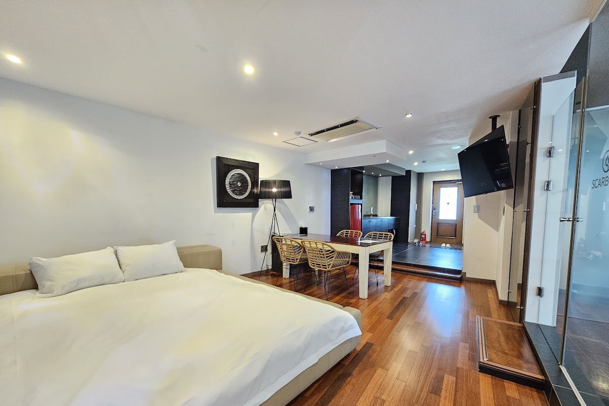 A modern hotel room with a large bed, a dining area with chairs, a flat-screen TV mounted on the wall, and a small kitchen area.