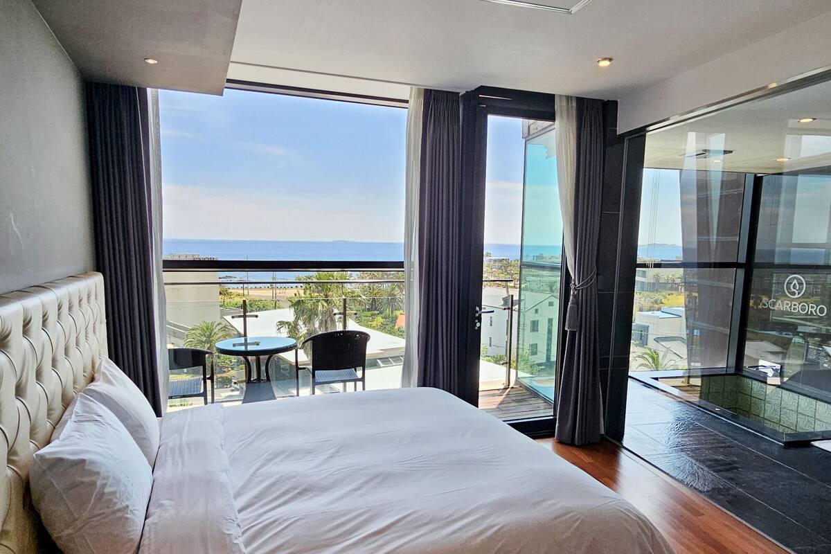 A modern hotel room with a large bed, a balcony with a view of the sea, and a glass door leading to an outdoor area.
