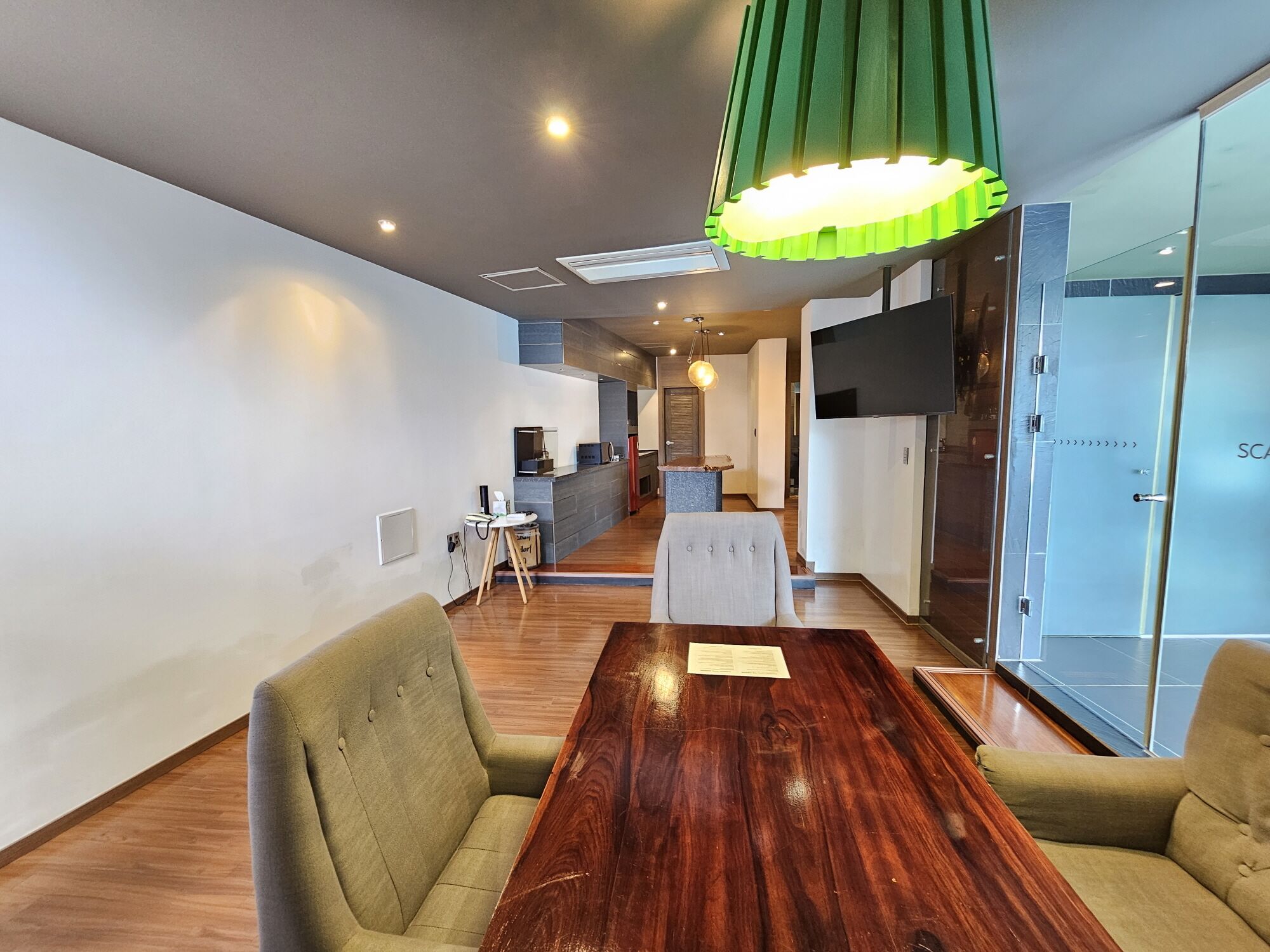 A modern office space with a wooden conference table, green pendant lighting, and a glass partition with a TV.