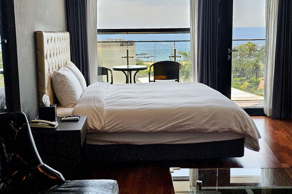A hotel room with a large bed, a bedside table with a lamp, a chair, and a small table. The room has a balcony with a view of the sea and buildings.