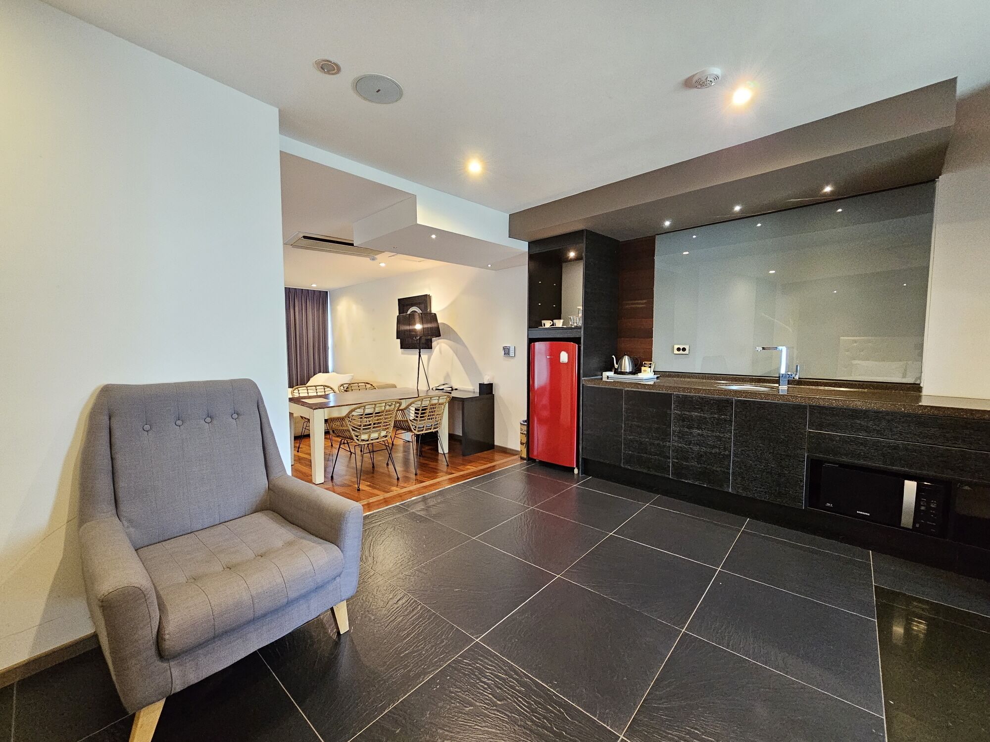 A modern living room with a grey sofa, a dining area with a table and chairs, and a kitchen with a red refrigerator and a black countertop.