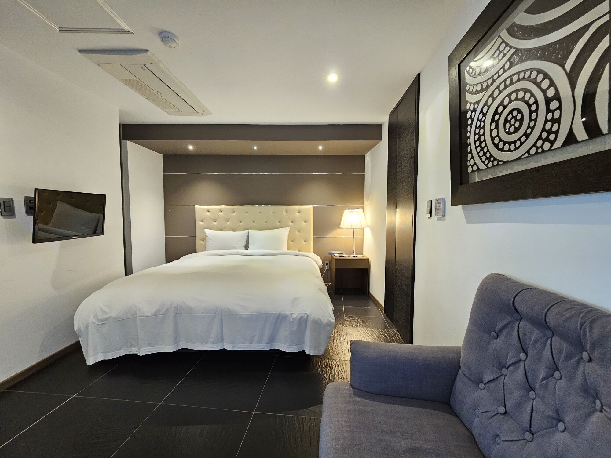 A modern hotel room with a large bed, a bedside table with a lamp, a sofa, and a decorative wall piece.