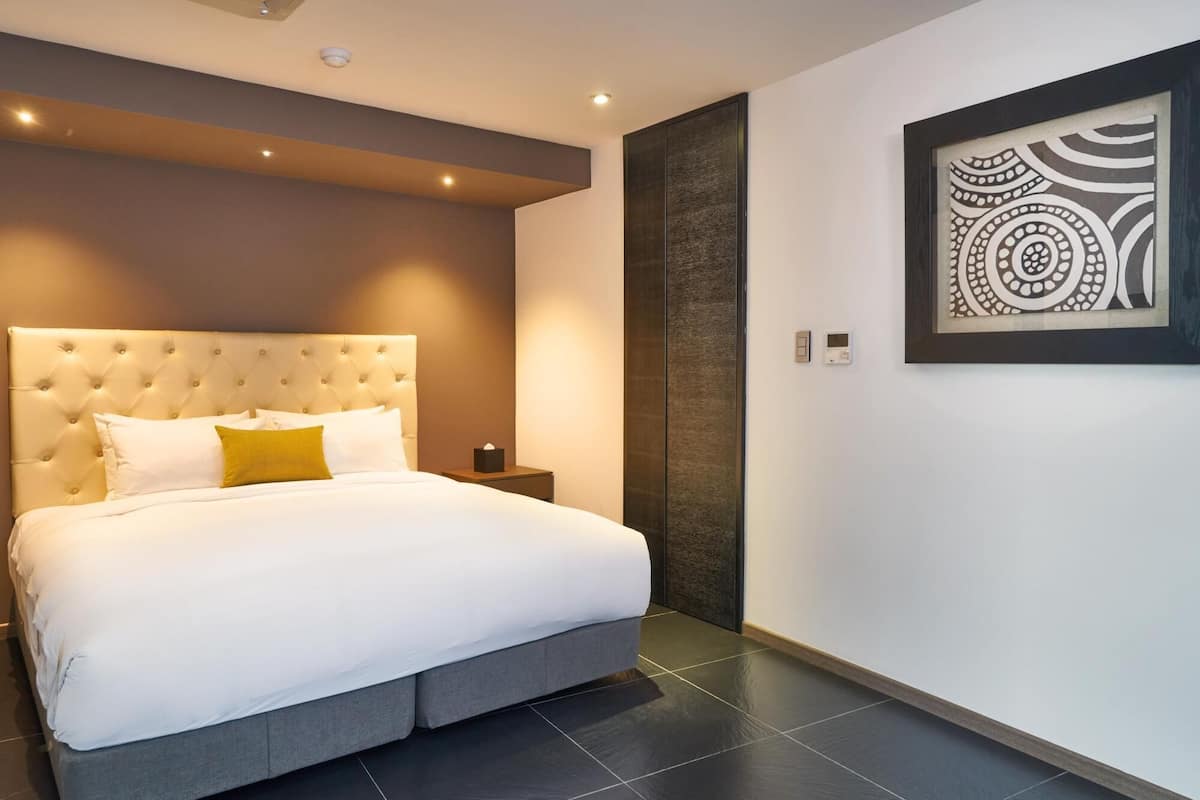 A modern hotel room with a tufted headboard, a large bed with white and yellow bedding, a nightstand, a wall-mounted artwork, and a dark tiled floor.