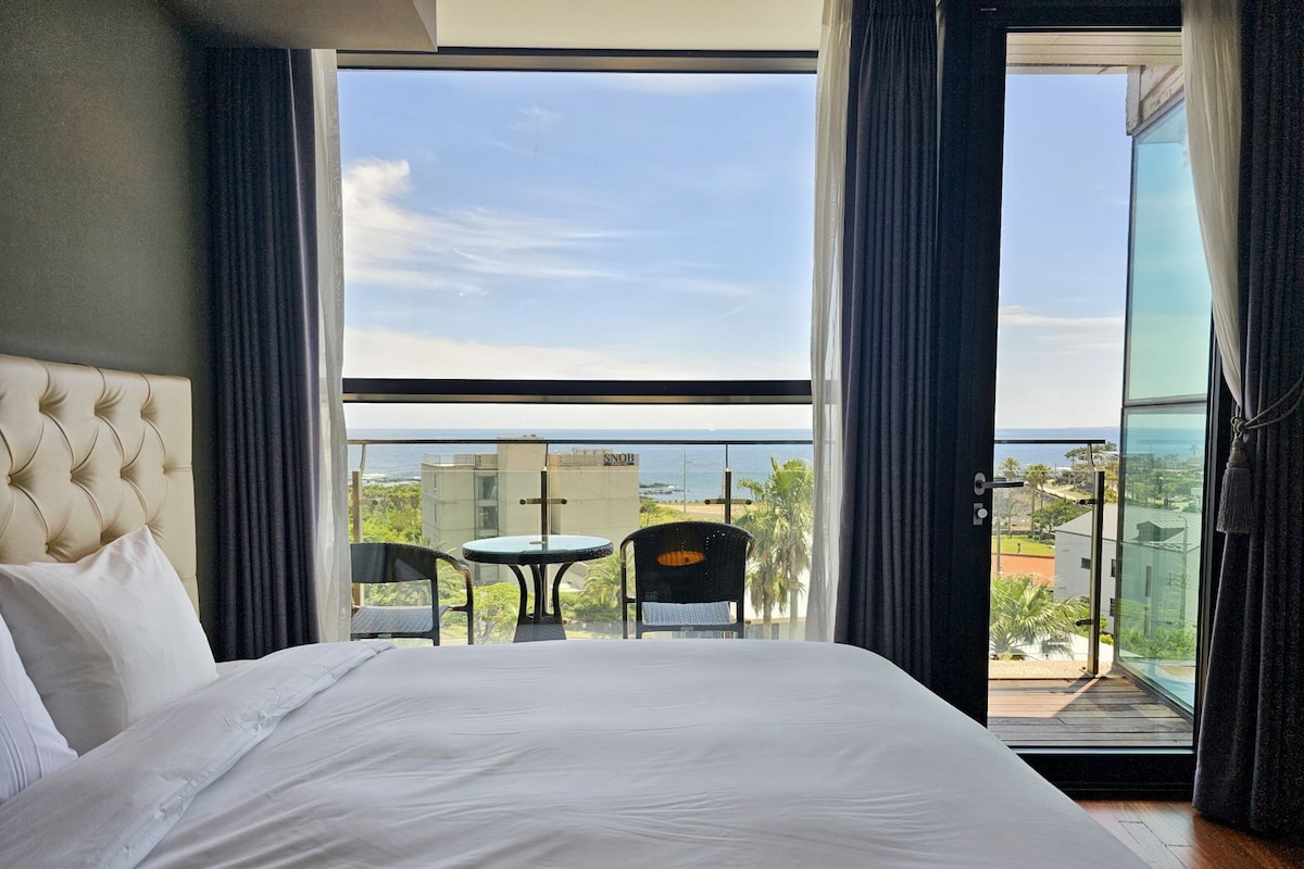 Habitación de hotel con una cama grande, un balcón con mesa y sillas, y vistas al océano.