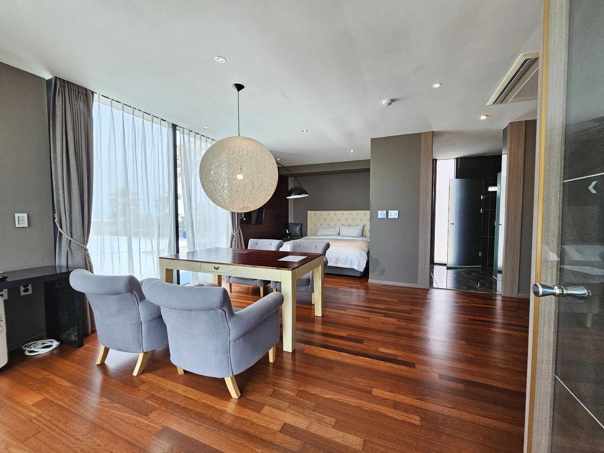 A modern hotel room with a wooden floor, a dining area with two grey chairs, a large round hanging light, and a bed with a headboard.