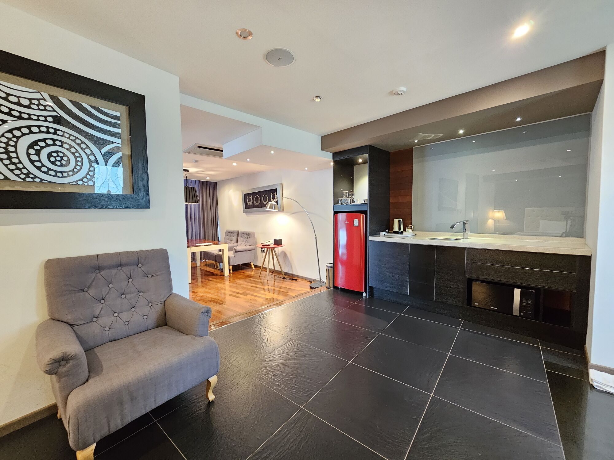 Een moderne woonkamer met een grijze bank, een eethoek met een tafel en stoelen, een keuken met een rode koelkast, en een grote spiegel boven de wasbak.