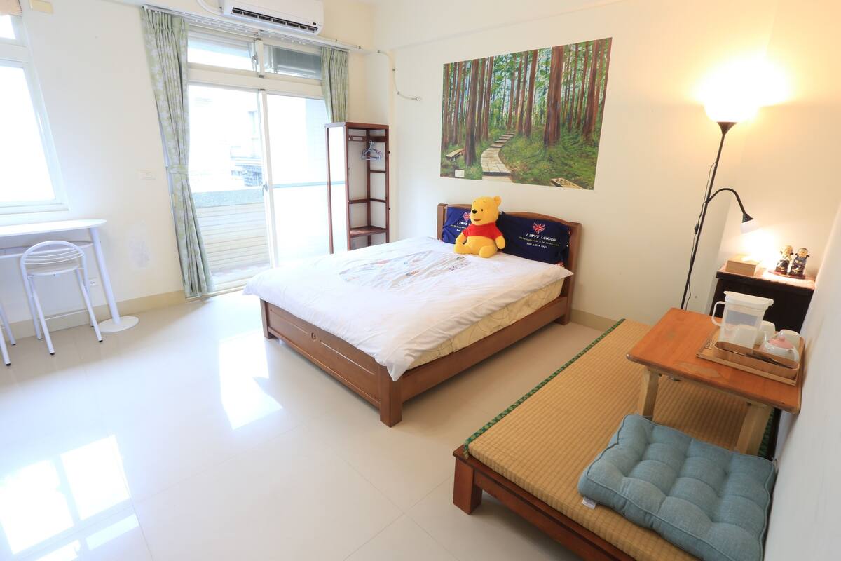 A bedroom with a bed, a desk, a chair, a bedside table with a lamp, a teddy bear, and a painting of a forest.