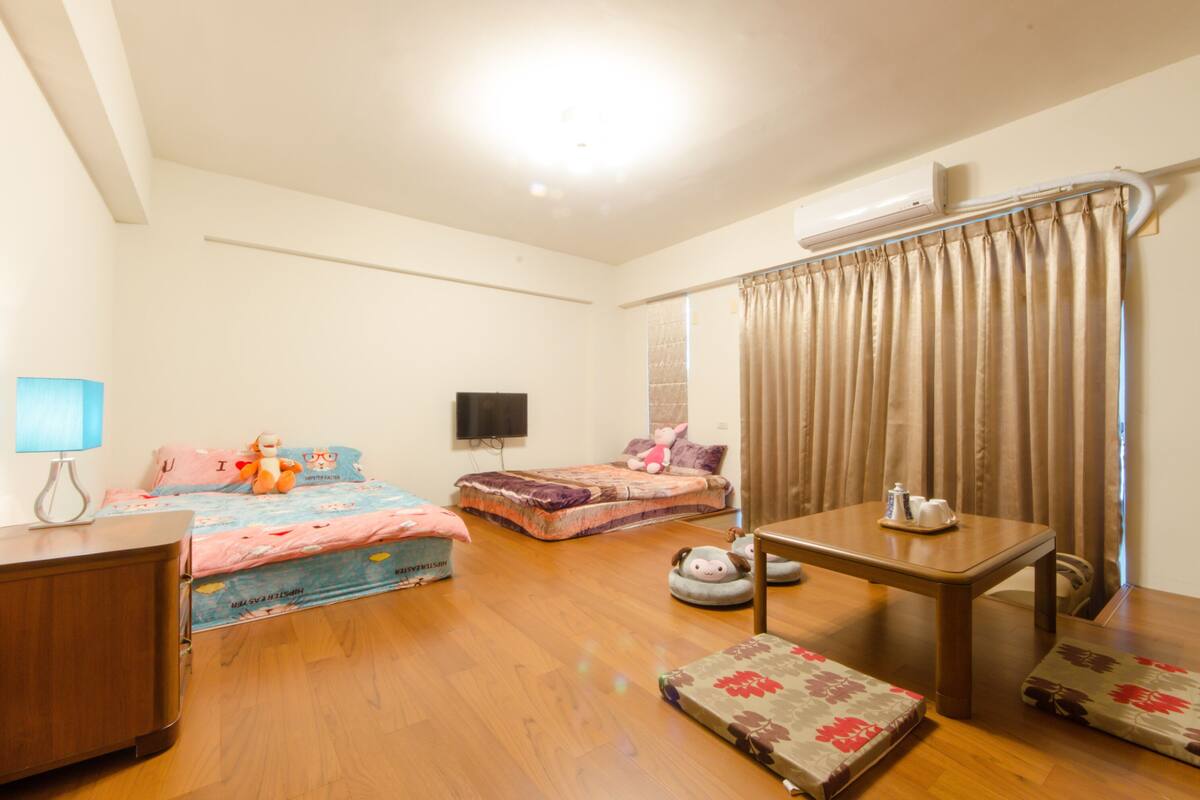 A bedroom with two beds, a desk, a chair, a TV, and a window with curtains.