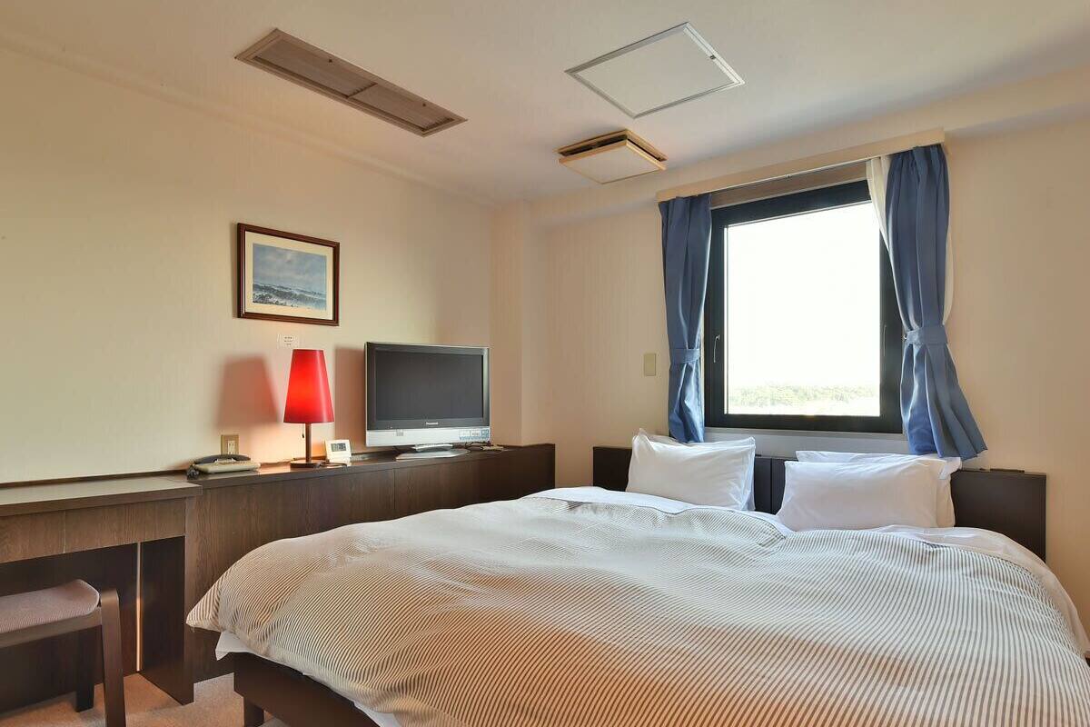 Ein Hotelzimmer mit einem großen Bett, einem Schreibtisch mit Computer, einem Fernseher und einem Fenster mit Vorhängen.