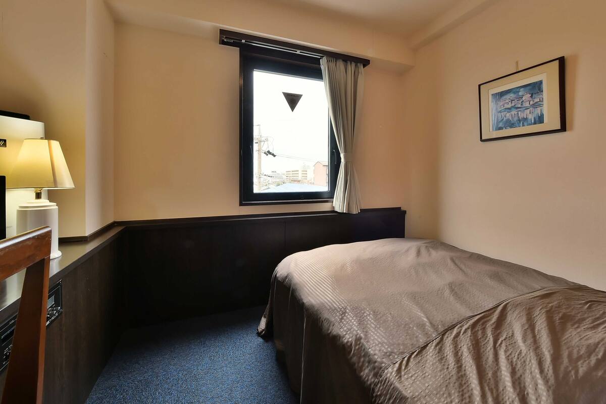 Ein Hotelzimmer mit einem Einzelbett, einem Schreibtisch mit Lampe, einem fenster mit Vorhängen und einem gerahmten Bild an der Wand.