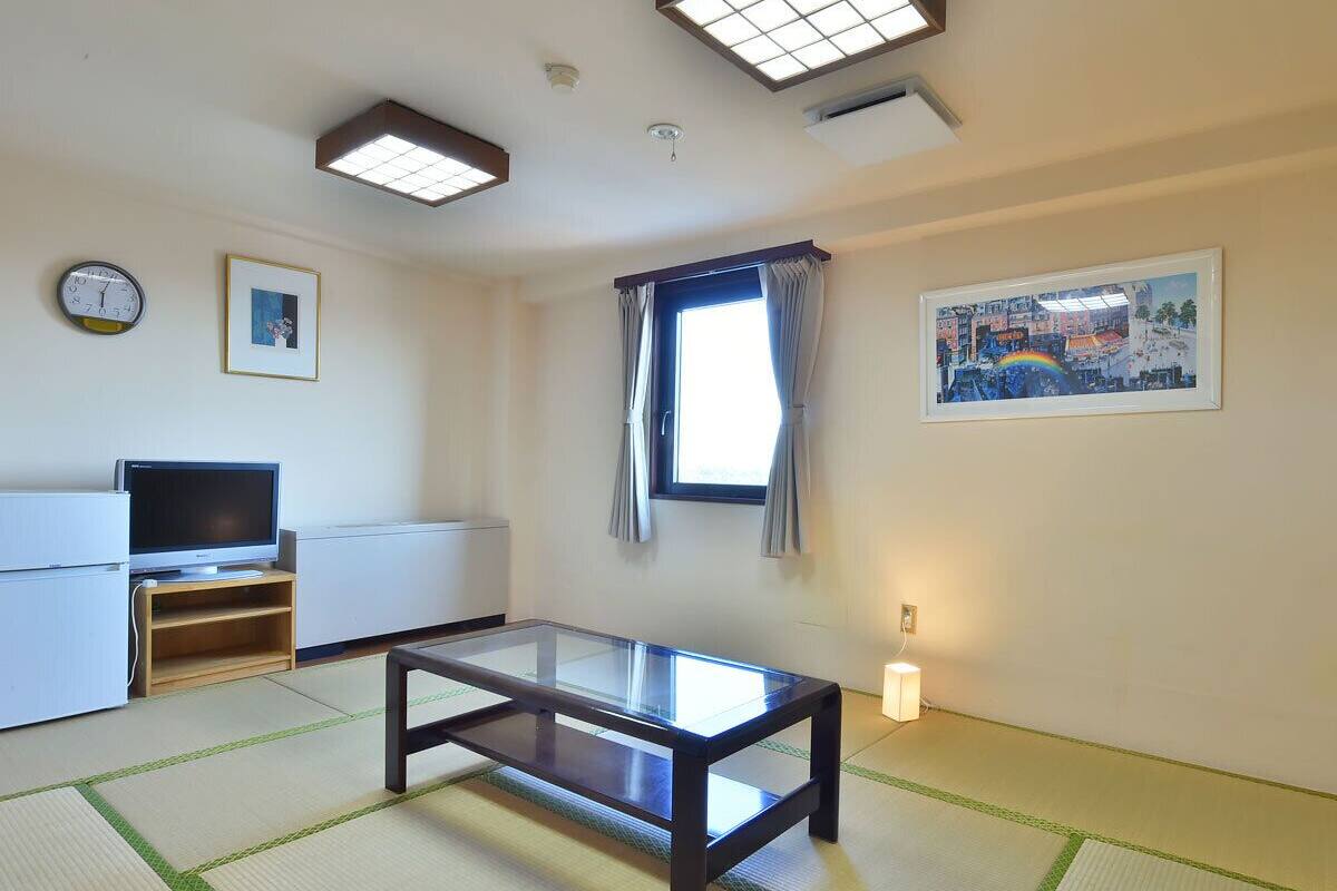 Ein Zimmer mit Tatami-Boden, einem Glastisch, einem Fernsehgerät auf einem Standfuß, einem Fenster mit Vorhängen und einem Bild an der Wand.