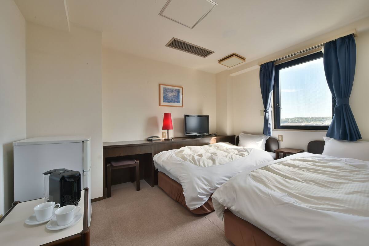 Ein Hotelzimmer mit zwei Betten, einem Schreibtisch mit Computer, einer Kaffeemaschine und einem Fenster mit Vorhängen.