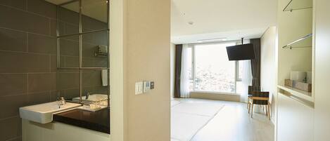 Un baño moderno con un espejo grande, un lavabo y una ducha. Hay un televisor montado en la pared y una ventana que proporciona luz natural.