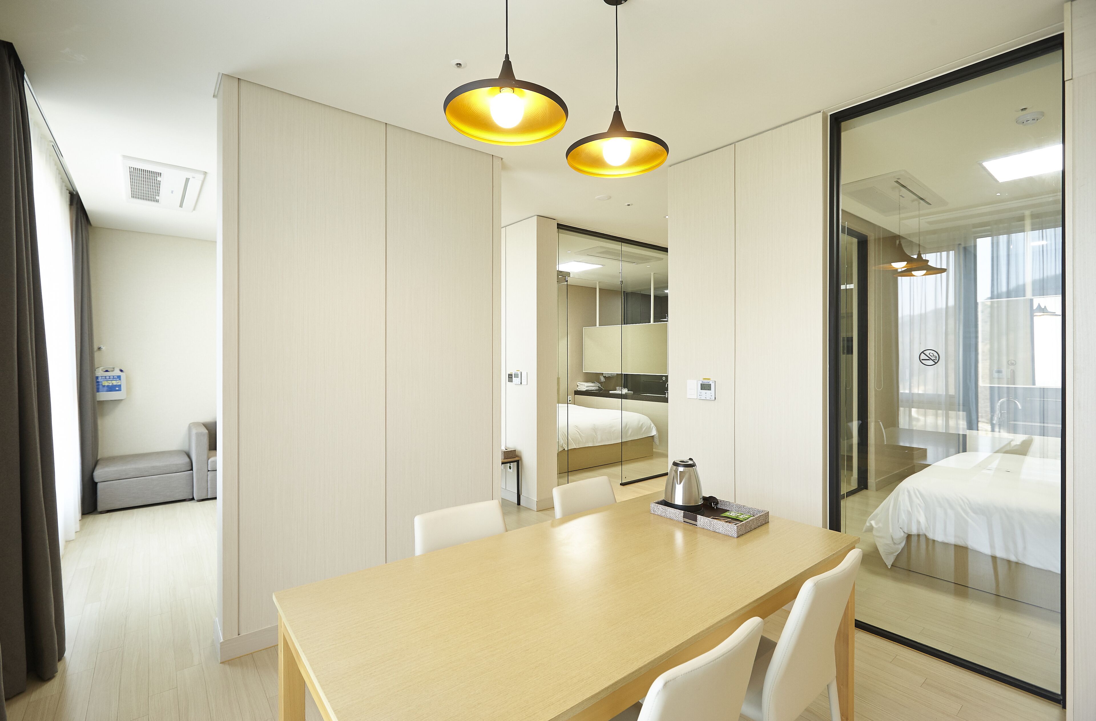 A hotel room with a wooden table, white chairs, a bed with white bedding, a glass partition, and a small kitchenette area.