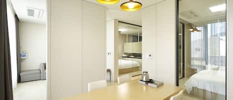 A hotel room with a wooden table, white chairs, a bed with white bedding, a glass partition, and a small kitchenette area.