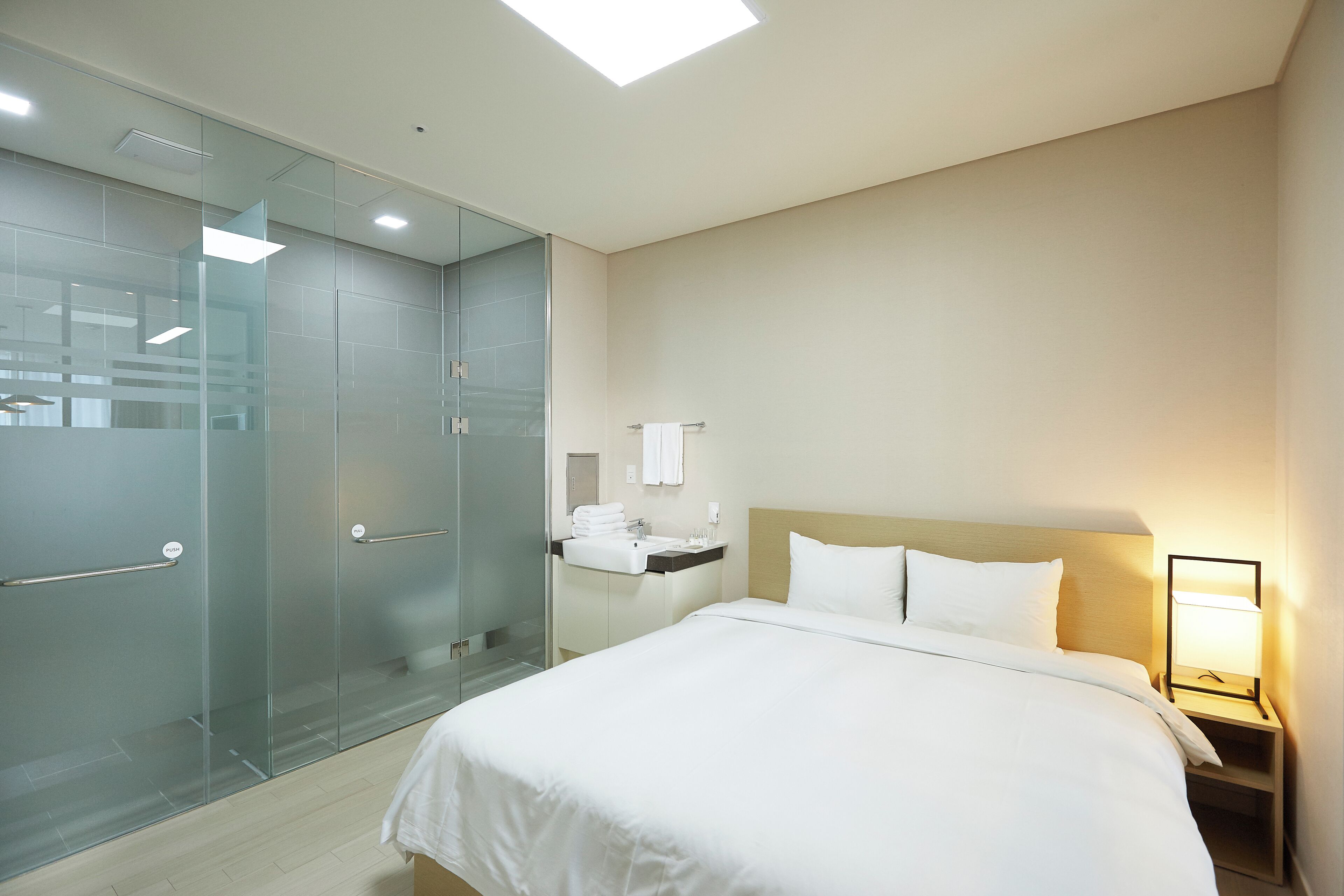 Una habitación de hotel con una cama grande, una mesita de noche con lámpara, ducha con mampara de cristal y un baño pequeño con lavabo y espejo.