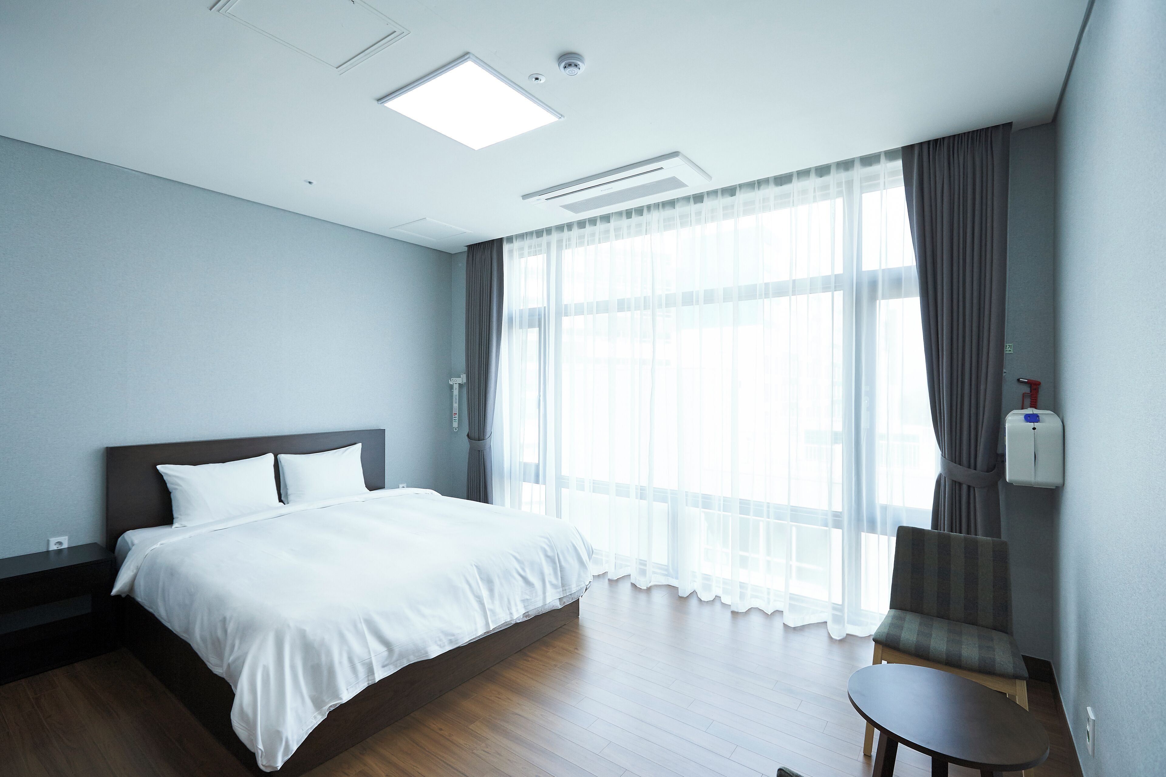 Una habitación de hotel con una cama grande, una mesita pequeña y una silla. Hay una ventana con cortinas y un aire acondicionado instalado en la pared.