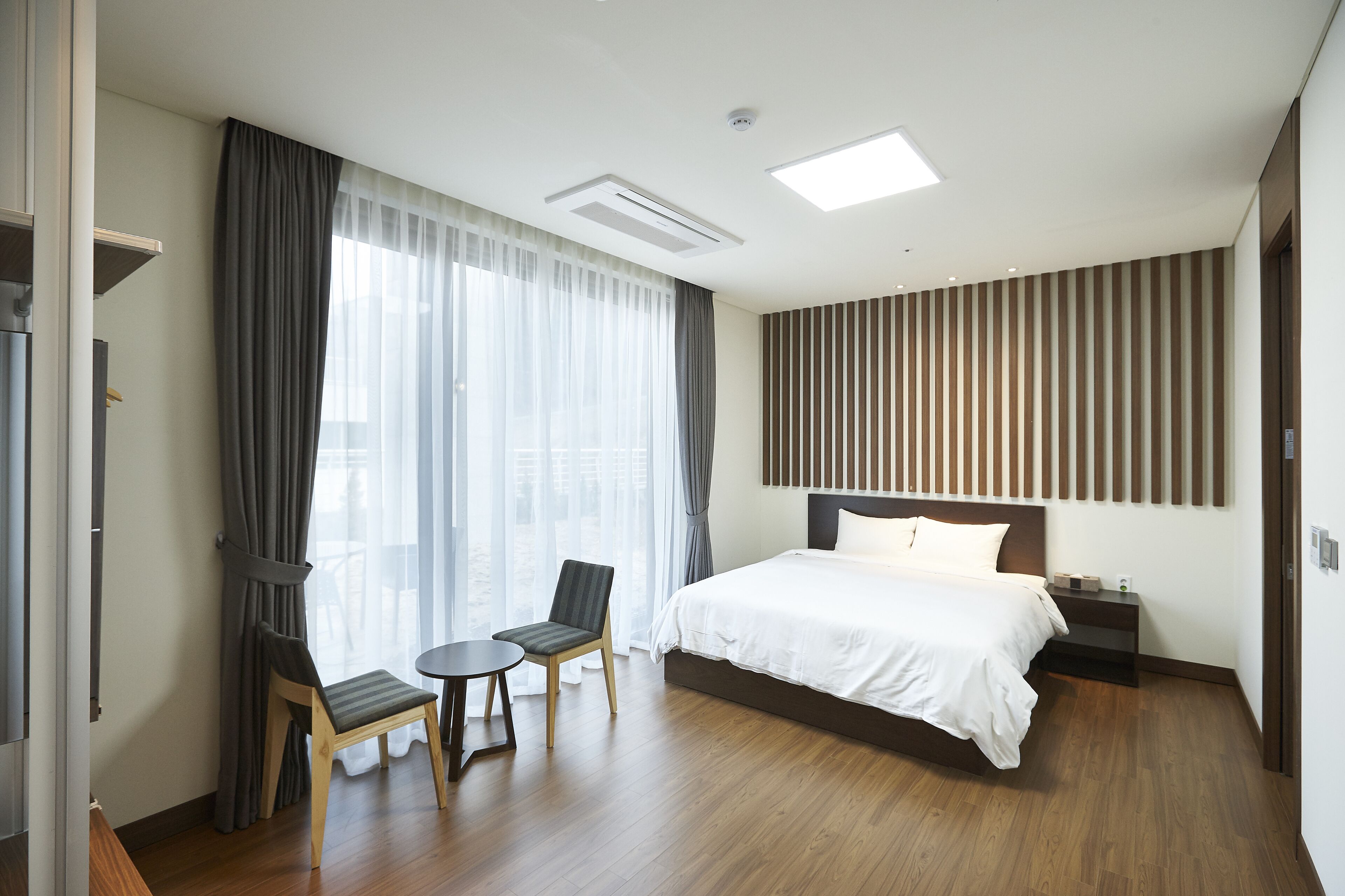 A modern hotel room with a large bed, a desk with two chairs, a view of the city through large windows, and a striped wall design.