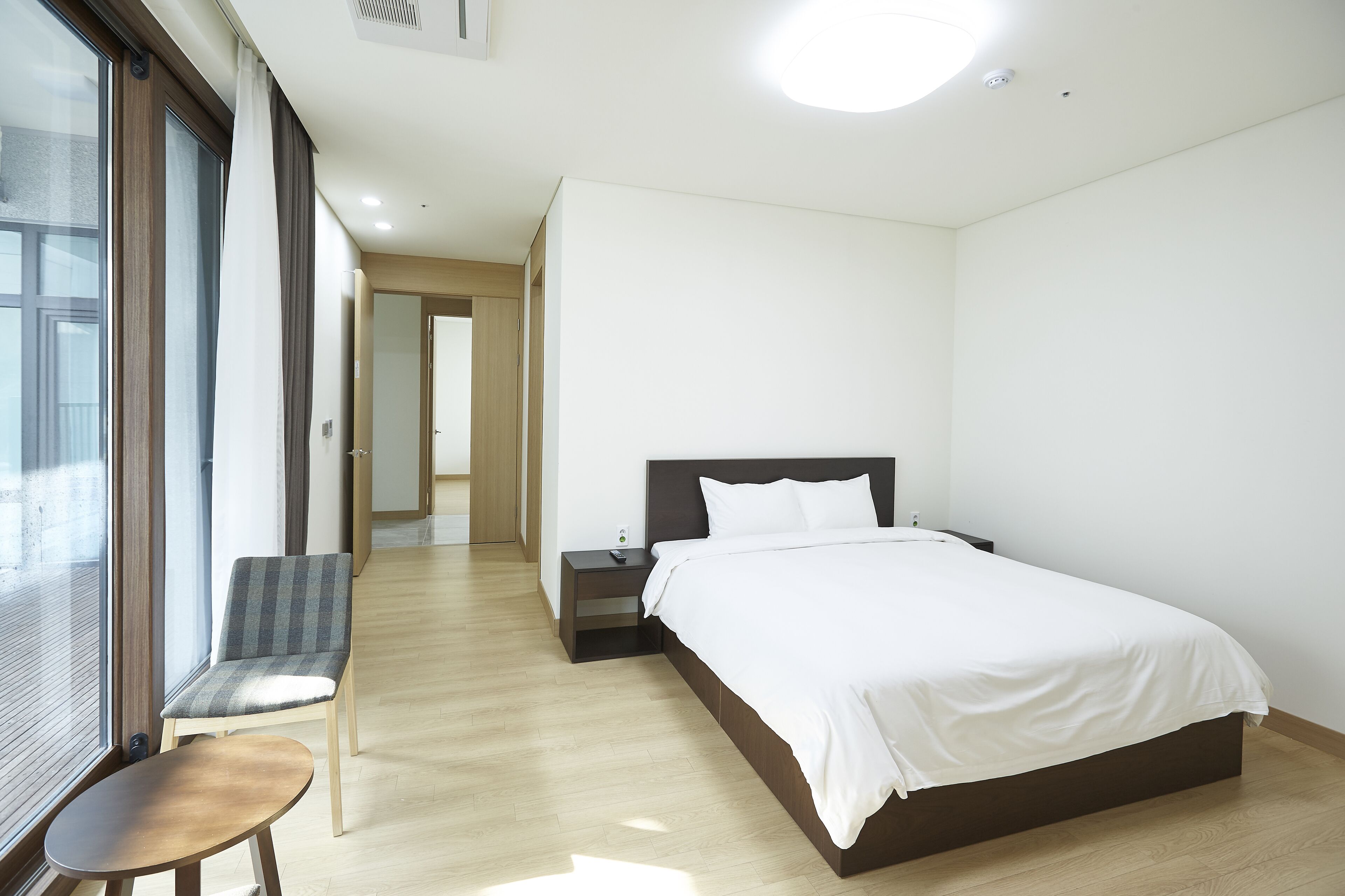 A modern hotel room with a large bed, a chair, a small table, and a view through a large window.