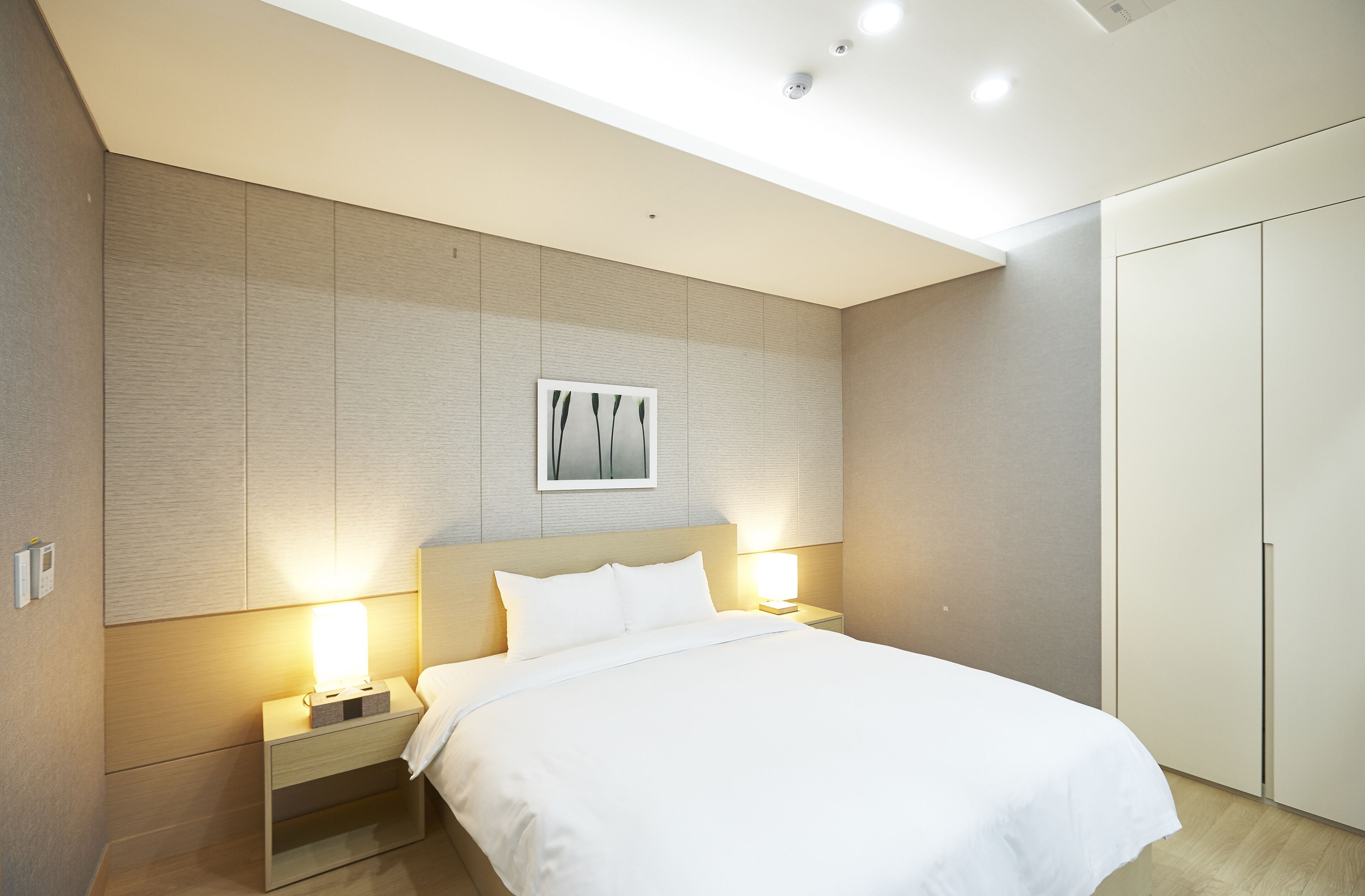 A modern hotel room with a large bed, bedside lamps, a framed artwork above the bed, and a wardrobe.