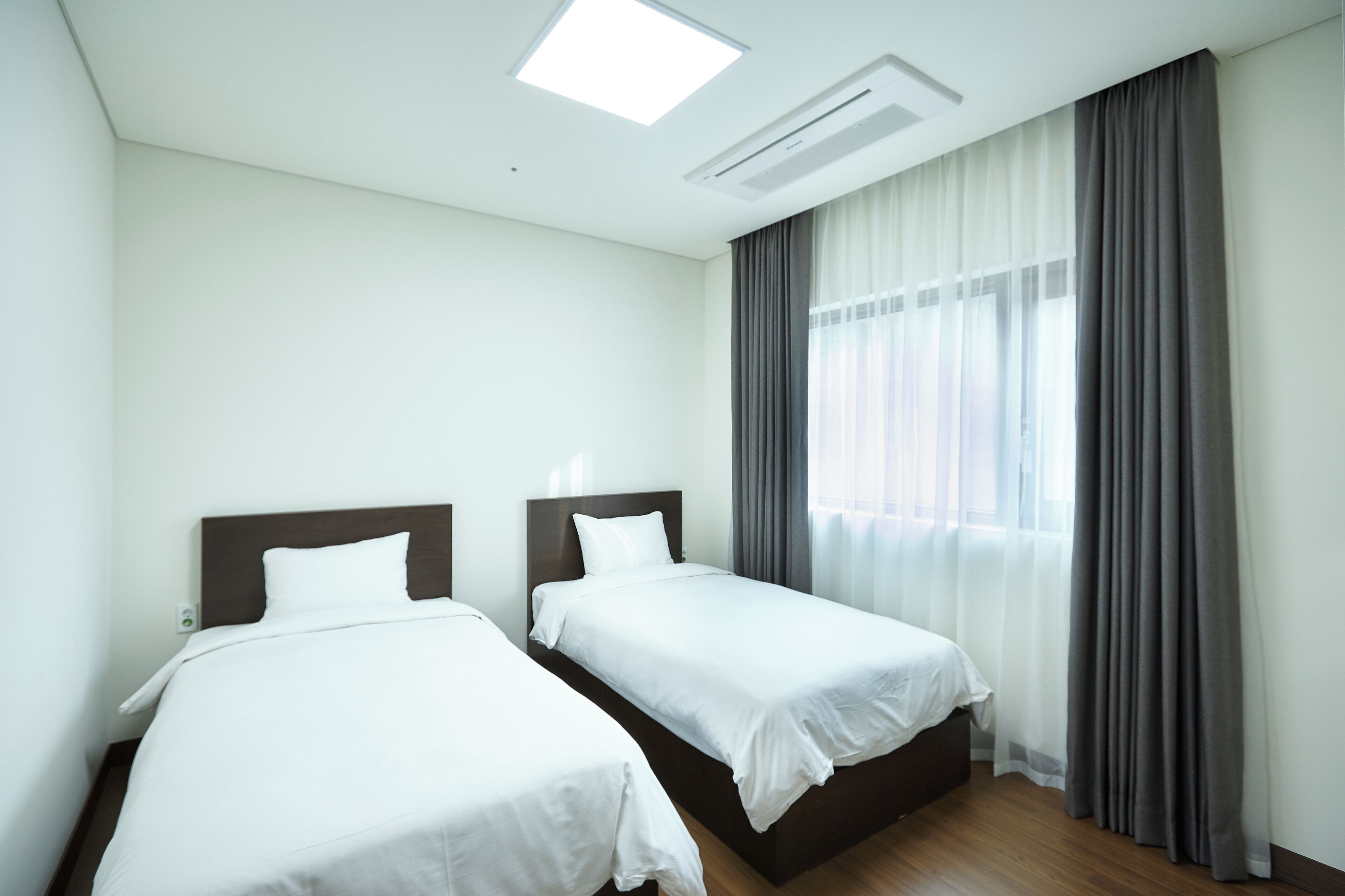 Ein Hotelzimmer mit zwei Einzelbetten, einem großen Fenster mit Vorhängen, einer Deckenleuchte und einer Wand-Klimaanlage.
