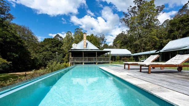 Una piscina con sillones de descanso, un porche cubierto y una casa con chimenea en un área boscosa.