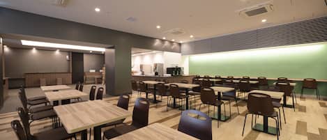 Une salle de classe moderne avec des tables et des chaises en bois, un tableau noir vert et, au fond, un coin cuisine.