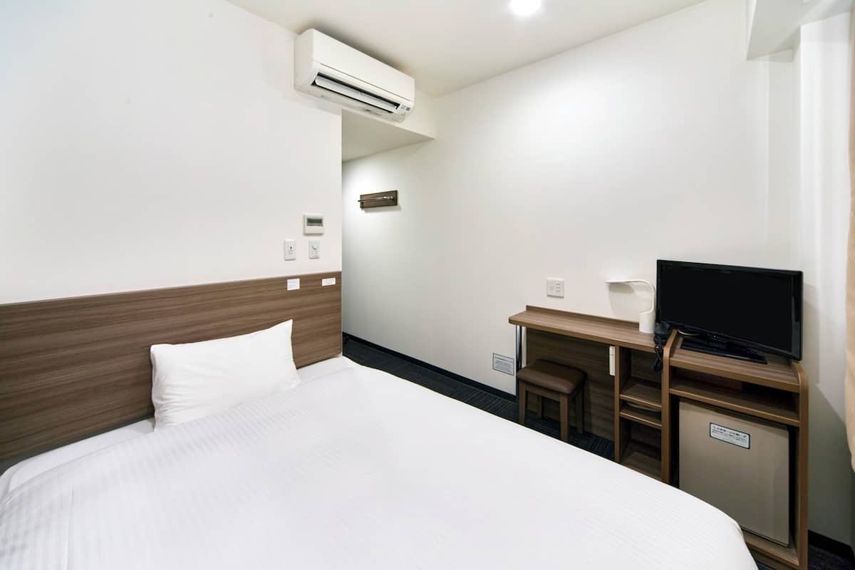 A hotel room with a bed, a desk with a television, and an air conditioning unit.