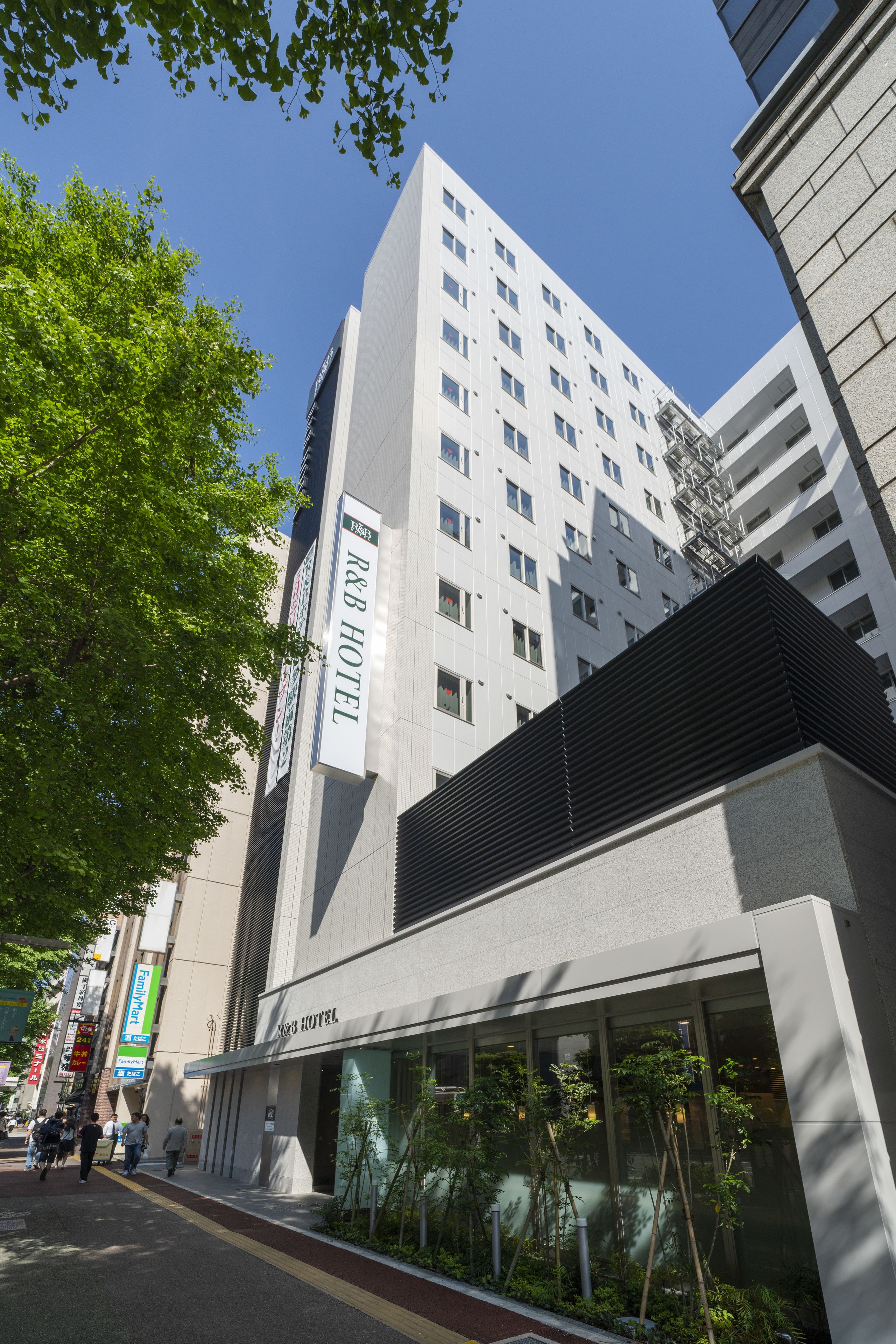Un moderno edificio de hotel con un llamativo letrero R&B Hotel, ventanas grandes y un espacio exterior con mesas y sillas.