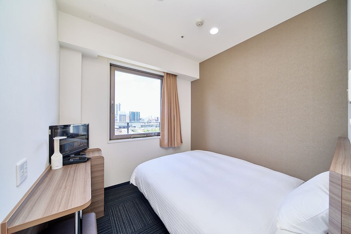 A hotel room with a bed, a desk with a television, a window with a city view, and a wall-mounted light.