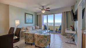 A hotel room with a balcony, a sofa, two armchairs, a glass coffee table, a TV, and a ceiling fan. - Ocean Front Delight - Ocean Walk (Daytona Beach)