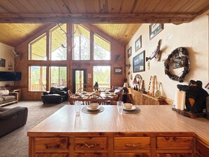 A rustic cabin interior with a dining area, wooden furniture, and large windows.