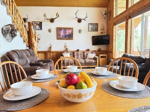 A dining area with a wooden table, chairs, a fruit bowl, and a staircase with antlers.