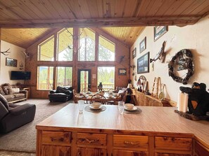 A rustic cabin interior with a large dining table, wooden walls, and a view of the outdoors.