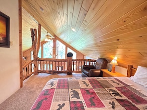 A bedroom with a wooden ceiling, a quilt with animal prints, a leather chair, and a nightstand with a lamp.