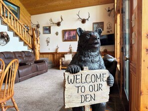 A bear holding a sign that reads 'WELCOME TO OUR DEN' in a cozy cabin interior with wooden walls, a leather couch, and antler decorations.