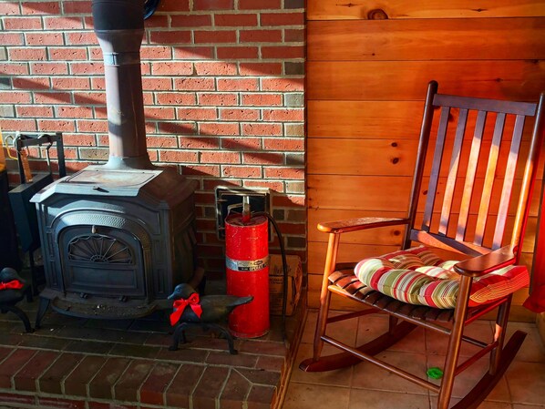 A rustic room with a brick wall, a wooden chair with a striped cushion, and a wood-burning stove. - 1800 sq ft♦ Kings Mountain Cottage ♦ On the Water (Orrington)
