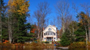 A two-story house with a prominent front entrance, surrounded by a forest and a lake.